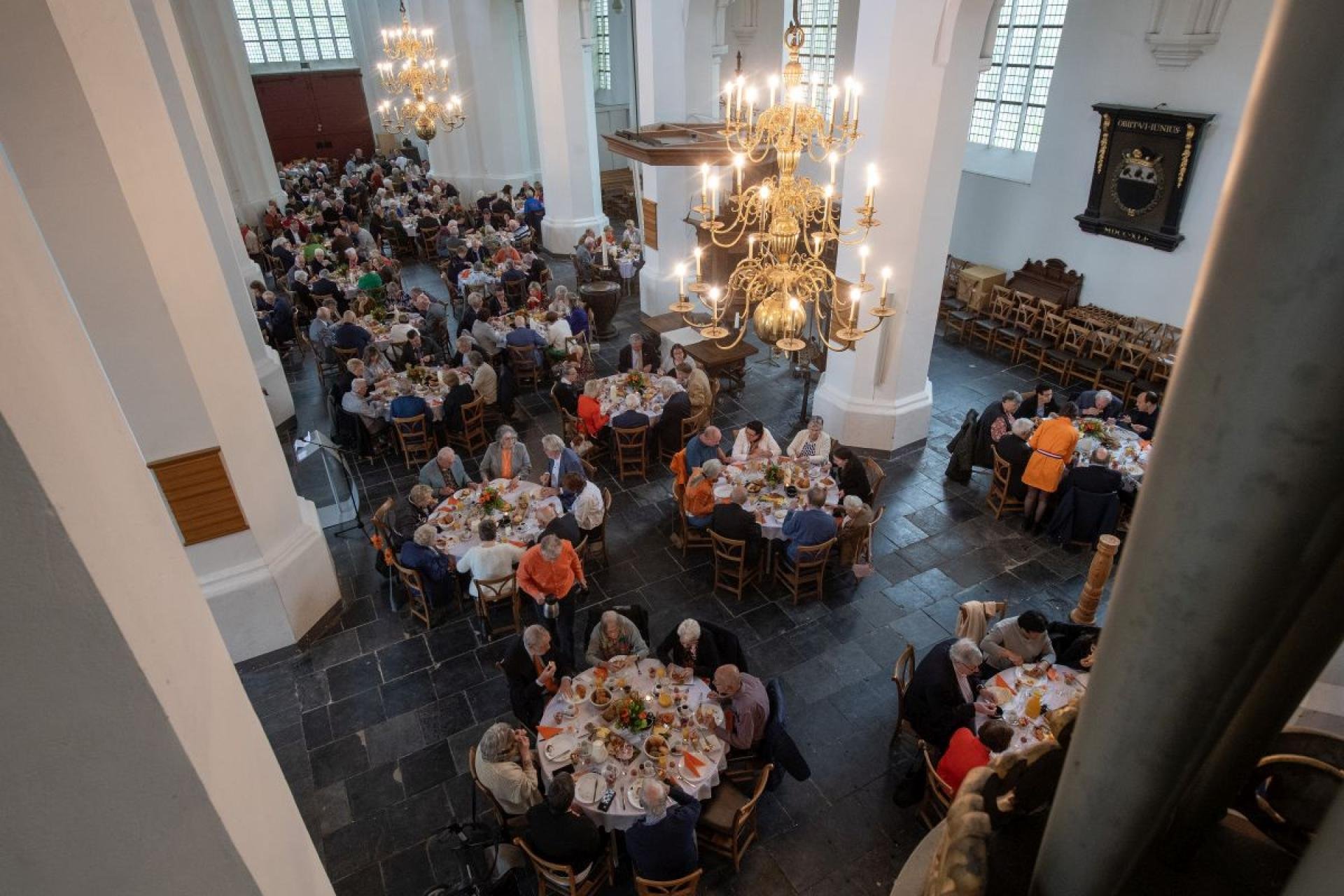 In een kerk staan heel veel ronde tafel waar mensen aan het ontbijten zijn. Foto is van bovenaf genomen