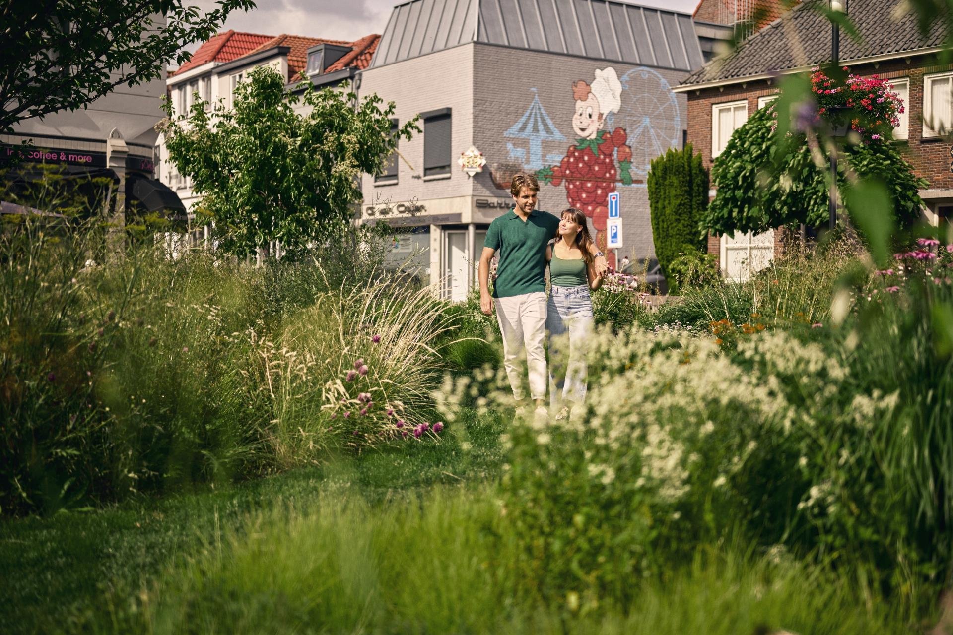 Man en vrouw lopen gearmd door een bloemrijke tuin in het centrum van Tiel. Op de achtergrond is er een grote tekening op de zijkant van een winkelpand te zien van Flipje