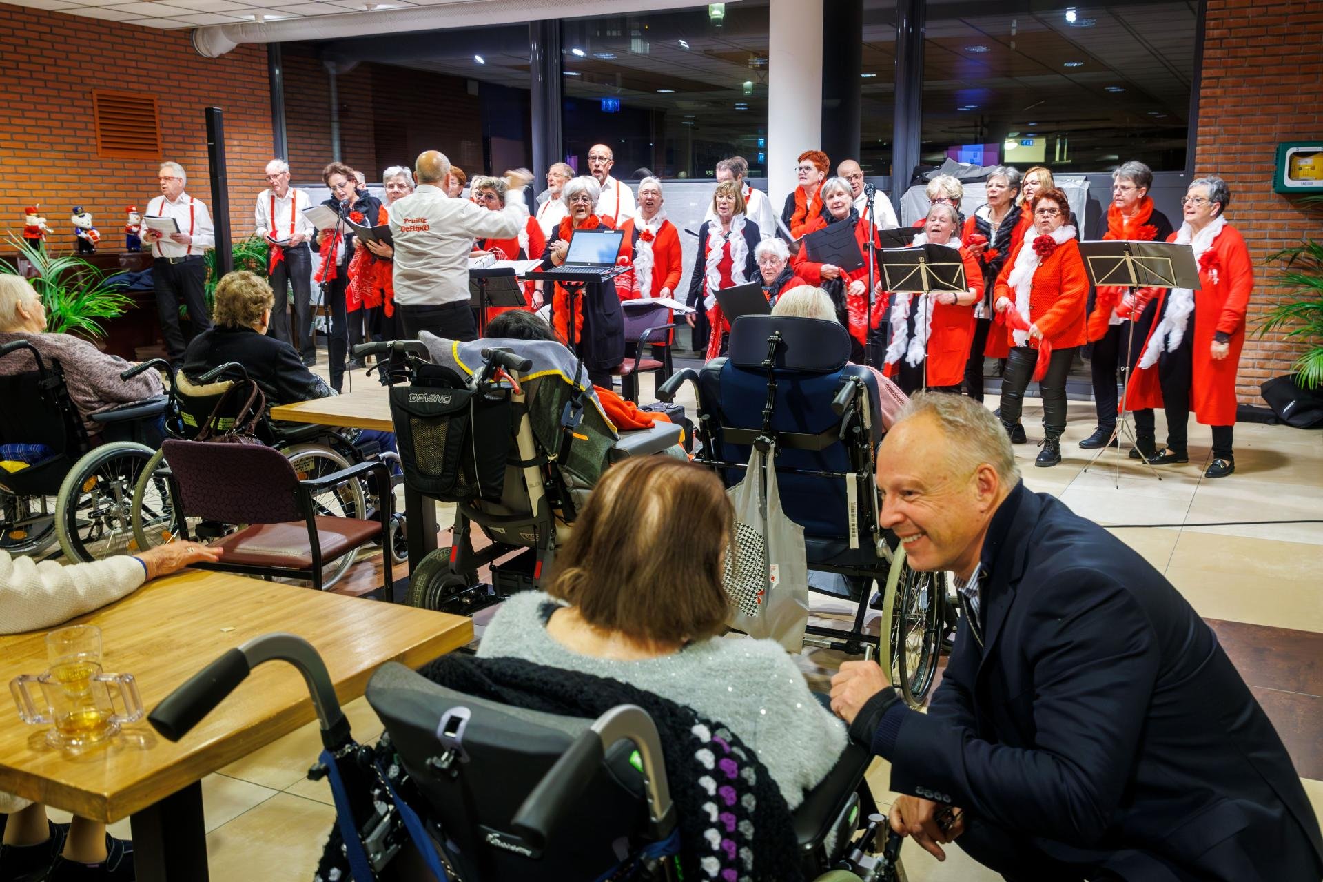 Op achtergrond koor in rood en wit. Oudere mensen in rolstoel luisteren op de rug gezien, behalve wethouder Dijkstra, hij luister met bewoner verzorgingstehuis mee