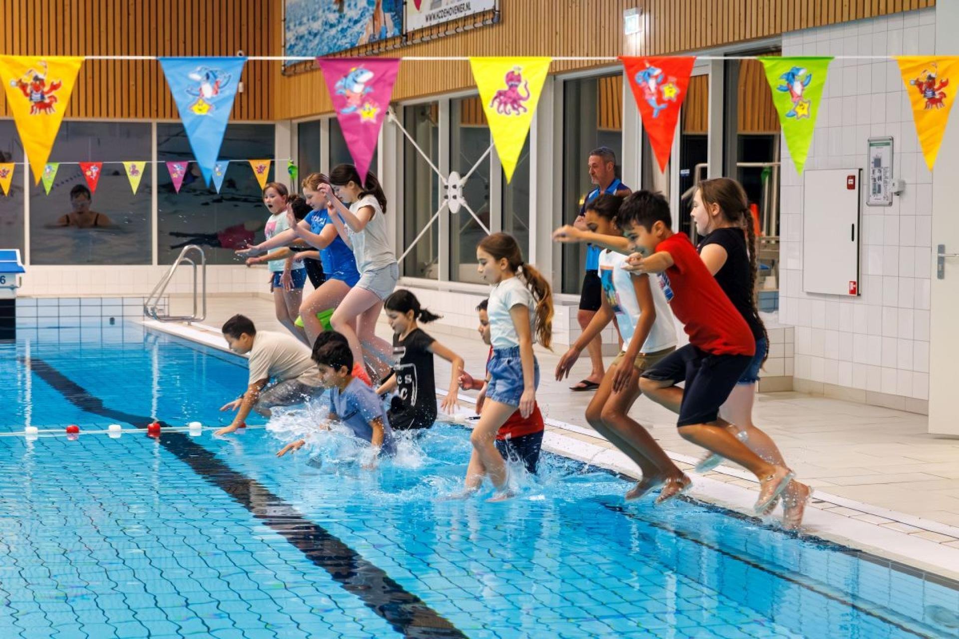 Kinderen in kleding spring in het water van een zwembad. Ook hangen er vlaggetjes in het zwembad, een slinger